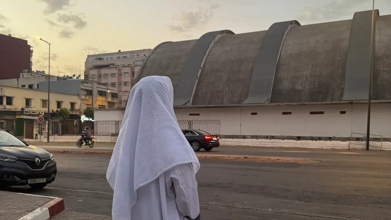 Une rue de Casablanca à la tombée du jour, vue depuis le trottoir pavé : une silhouette de dos en djellaba blanche, la halle du marché aux toits arrondis en face, et un ciel encore doré.